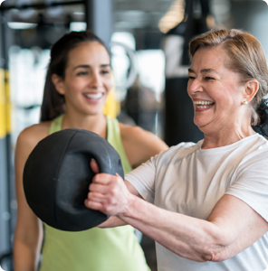 alt="Smiling senior woman lifting a medicine ball during a workout with a female personal trainer smiling in the background."