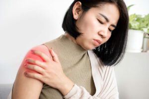 A woman is experiencing pain and stiffness in her shoulder due to frozen shoulder, and she is holding her shoulder for relief.
