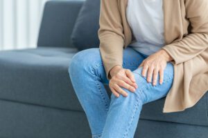 A woman suffering from knee pain due to osteoarthritis is sitting on the sofa in her living room while experiencing the pain.