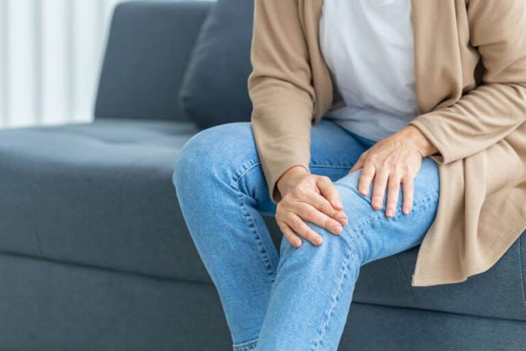 A woman suffering from knee pain due to osteoarthritis is sitting on the sofa in her living room while experiencing the pain.