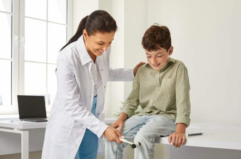 A pediatric orthopedic doctor examining a happy little child and using a hammer to check his knee reflex.